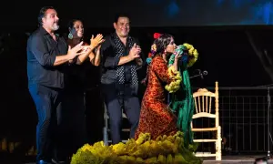 Una bailaora de flamenco andalusi, con un vestido rojo y amarillo, besa un mantón verde. A su lado, tres músicos aplauden.