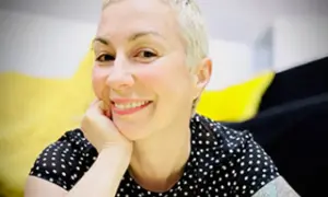 Laura Veltz, a blonde woman with short hair, smiles and rests her chin on her hand. She wears a black polka dot shirt.