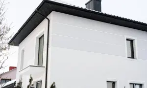 Nowoczesny dom kostka z jasną elewacją, czarnym dachem i kominem. Śnieg na dachu i gałęziach drzew.