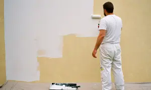 Un peintre en blanc est en train d'enduire sur peinture une partie d'un mur extérieur jaune avec un rouleau blanc.