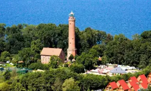 Latarnia morska Sarbinowo góruje nad zielonymi drzewami, z błękitnym morzem w tle. Obok niej budynki i czerwone dachy domków letniskowych.