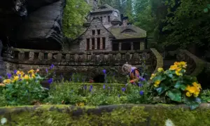 Kamienny domek w skalnym otoczeniu, idealne miejsce na czeska szwajcaria noclegi. Wokół kwitnące kwiaty i zielone drzewa.