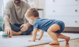 Dziecko w niebieskim body raczkuje na dywanie, obok siedzi ojciec.