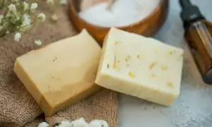 Dwa kostki mydła, jedno jasne, drugie z widocznymi drobinkami, leżą na brązowym ręczniku. W tle miska z solą i buteleczka z olejkiem. Mydło siarkowe właściwości doceniane w pielęgnacji.