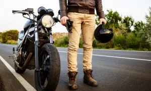 Motocyklista w skórzanej kurtce i beżowych spodniach trzyma kask i rękawice, stojąc obok motocykla na drodze.