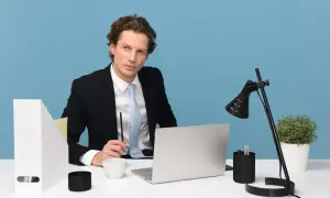 Młody manager w garniturze siedzi przy biurku, trzymając długopis. Na biurku laptop, filiżanka i lampka.