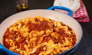 Breite Tagliatelle mit herzhaftem Ragu Bolognese in einer blauen Pfanne. Daneben ein Glas Olivenöl und ein kariertes Tuch.
