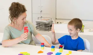 Kobieta pokazuje chłopcu liczbę 5, podczas gdy on podnosi rękę. To scena z zajęć surdopedagogiki, gdzie nauka odbywa się przez zabawę.