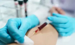 Felczer w niebieskich rękawiczkach pobiera krew do strzykawki z ręki pacjenta. W tle probówki.