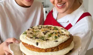 Due persone sorridono mostrando una torta a strati decorata con glassa, semi di zucca e scorza d'arancia. La ricetta della carrot cake è un successo!