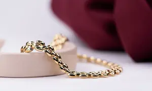Pulsera de eslabones ovalados en oro, brillando sobre un soporte beige, con un fondo de tela burdeos.