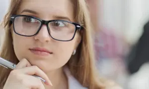 Młoda kobieta w okularach, z długopisem w dłoni, zamyślona. Jej słaby wzrok nie przeszkadza jej w pracy.
