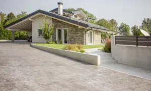 Un moderno **pavimento in cemento per esterno** accoglie una casa con tetto spiovente e rivestimento in pietra.