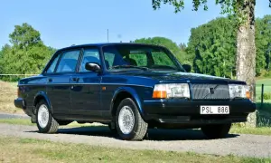Mörkblå Volvo 240 från 1993 parkerad på en grusväg med grönskande träd och fält i bakgrunden.