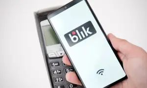 Dłoń trzyma smartfon z logo Blik nad terminalem płatniczym. Szybki przelew na telefon bez obaw o limit.