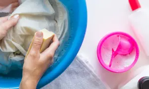 Hände reiben mit Kernseife über ein Kleidungsstück in einer blauen Schüssel, um fettflecken aus kleidung entfernen. Waschmittel und Flaschen daneben.