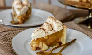 Ein Stück Apfelkuchen mit Sahne, bestreut mit Zimt, auf einem Teller mit goldenen Löffeln.
