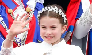 Uśmiechnięta księżniczka Charlotte w tiarze macha ręką, ubrana w białą sukienkę i ozdobne szaty.