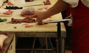 Rzeźnik w czerwonym fartuchu kroi mięso na desce. W tle widać kawałki mięsa i opakowania.
