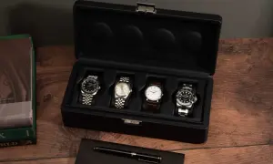 Una caja de reloj negra abierta muestra cuatro relojes de lujo. A la izquierda, libros de Rolex y un bolígrafo.