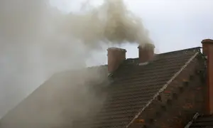 Gęsty, żółty dym z komina unosi się nad brązowym dachem.