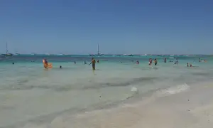 Słoneczny dzień na plaży Es Trenc. Ludzie kąpią się w turkusowej wodzie, a na horyzoncie widać żaglówki.