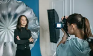 Dzień fotografa w studiu. Młoda kobieta z aparatem w ręku, w tle modelka pozująca na tle niebieskiego tła i srebrnego parasola.