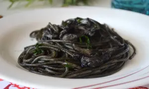 Spaghetti al nero di seppia, un piatto intenso e saporito, servito su un piatto bianco con guarnizioni verdi.