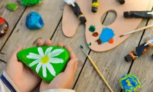 Kreative Ideen zum Steine bemalen für Kinder: Ein grüner Stein mit Gänseblümchen, bunte Farben und Pinsel laden zum Malen ein.