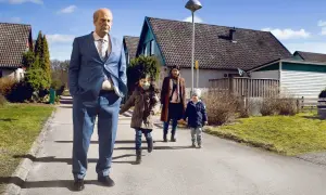 En man i kostym går med en familj i ett villaområde, en scen som kan passa i svenska komedier.