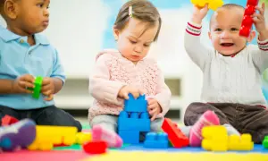 Three toddlers engage in parallel play, each absorbed in their own colorful blocks. One stacks blue blocks, another holds red and yellow toys, and the third plays with green.