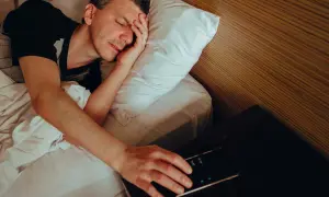 Man in bed, hand reaching for phone alarm. Does snoozing make you more tired?