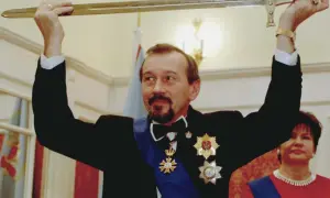 Mężczyzna w czarnym garniturze i orderach trzyma miecz nad głową. Obok kobieta w czerwonej sukni.