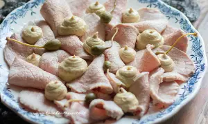 Vitello tonnato della nonna: fette sottili di carne rosa, guarnite con salsa tonnata e capperi, su un piatto bianco con decori blu.