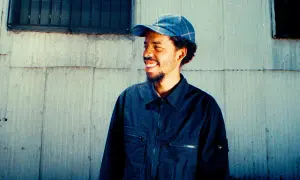 Earl Sweatshirt, smiling and wearing a blue plaid cap and dark jacket, stands against a weathered wall. He's ready to drop his next earl sweatshirt setlist.