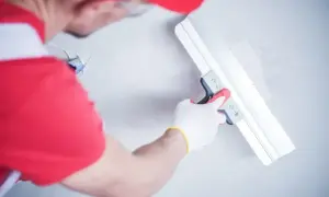 Gipsar w czerwonej czapce i rękawiczkach wygładza ścianę gładzią za pomocą szerokiej pacy.