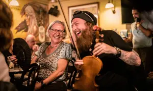 Una mujer ríe mientras un violinista con barba toca. El ambiente es festivo, ¡qué buen craic!