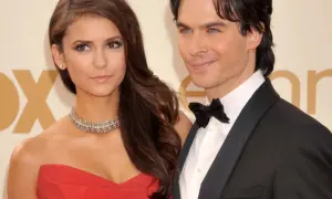 Nina Dobrev w czerwonej sukni i jej partner w smokingu, uśmiechnięci na ściance.