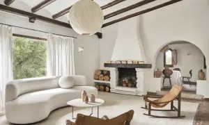 Salón acogedor con sofá curvo blanco, chimenea de piedra y mecedora de cuero. Un ejemplo de casas de campo sencillas y bonitas.