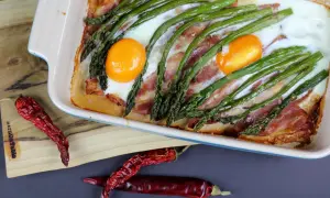 Pyszne śniadanie ze szparagami, jajkami i bekonem zapieczone w naczyniu. Obok leżą suszone papryczki chili.