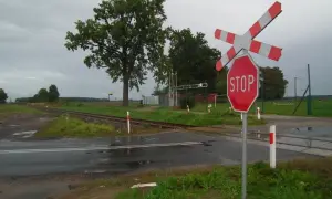 Przejazd kolejowy z zaporami i znakiem STOP. Torowisko przecina mokrą drogę, a w tle widać drzewa i boisko.