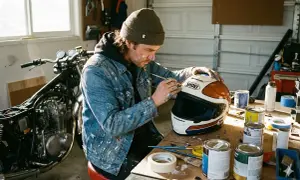 Mężczyzna w dżinsowej kurtce z plamami farby tworzy unikalny wzór na białym kasku motocyklowym.
