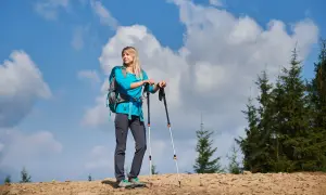 Kobieta z plecakiem i kijkami trekkingowymi na górskim szlaku. Zastanawia się, jak przymocować kijki do plecaka, by mieć wolne ręce.
