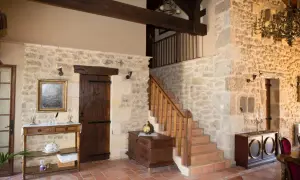 Interior de una casa antigua con paredes de piedra, escaleras de madera, un baúl y muebles rústicos.