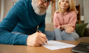 Starszy mężczyzna z brodą podpisuje dokumenty, podczas gdy kobieta siedzi obok. Rozmowa o dziedziczeniu.
