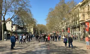 Tłum ludzi spaceruje po słynnej alei La Rambla w Barcelonie, otoczonej drzewami i zabytkowymi budynkami.