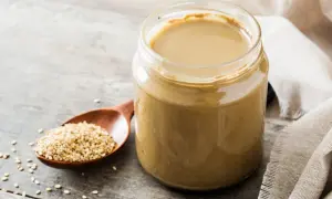 Słoik z gładkim, kremowym tahini obok drewnianej łyżki pełnej nasion sezamu. To właśnie tahini co to jest, czyli pasta z sezamu.
