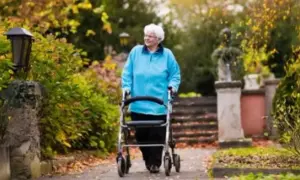 Elderly woman in blue jacket walks with a walker in a park with autumn foliage.