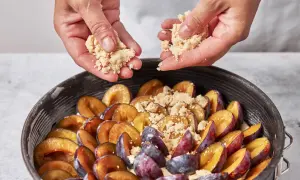 Hände streuen Streusel über saftige Pflaumen in einer Springform. Der pflaumenkuchen nimmt Gestalt an.