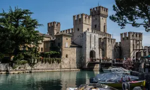 Il castello di Moniga si affaccia sull'acqua, con torri merlate e un ponte pedonale affollato. Barche ormeggiate completano la scena.